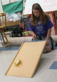 A young person kneels on the floor near a ramp made from a propped-up (angled) piece of wood. They have released a small wheeled vehicle from the top of the ramp, which rolls down the ramp towards the linoleum floor.