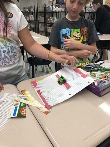 Two students test elements of friction using a green toy car, and an inclined ramp set up on a textbook.